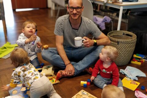 Mies istumassa lattialla kahvikuppi kädessä, lapsia ympärillä