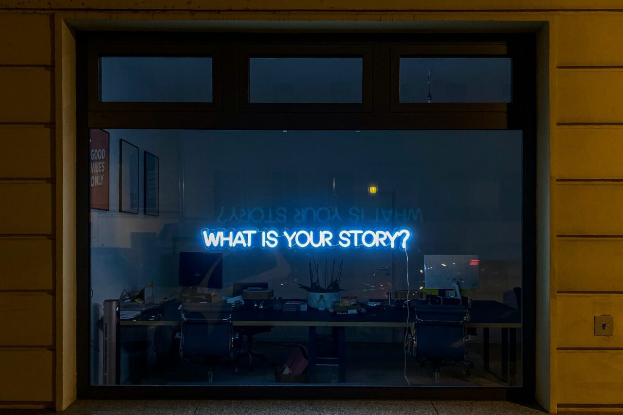 Pimeänä olevan liiketilan ikkuna, johon heijastettu sinisellä teksti What is your story?