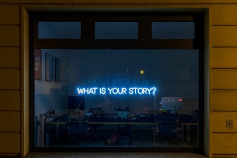 Pimeänä olevan liiketilan ikkuna, johon heijastettu sinisellä teksti What is your story?
