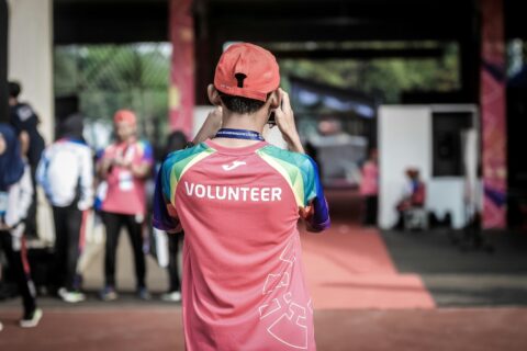 Nuori mies kuvattuna takaapäin valokuvaamassa taustalla olevia ihmisiä ja taphtumia. Miehen punaisessa teepaidassa lukee valkoisella VOLUNTEER