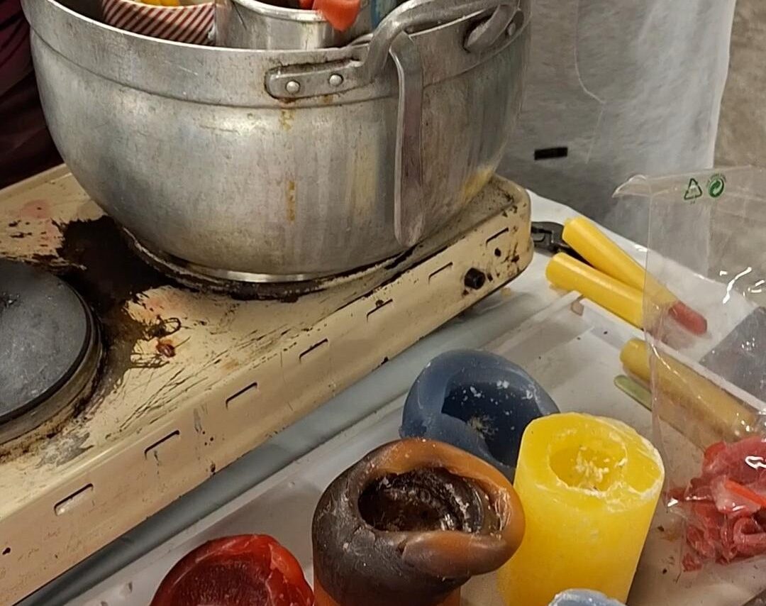 Puoliksi poltettuja kynttilöitä etualalla, taustalla liedellä vanha teräskattila, missä sulamassa kynttilänvahaa