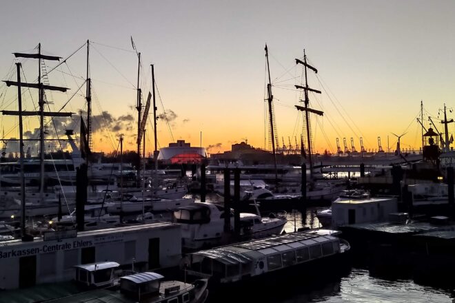 Auringonnousu talvisessa satamassa Hampurissa. Aurinko pilkottaa taustalla, edessä huurteisia laivoja