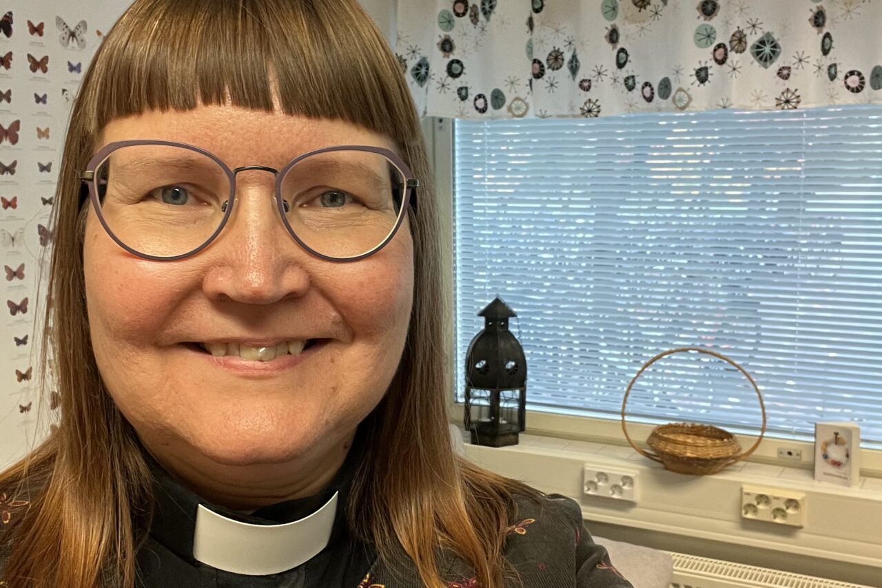 Kameraan hymyilevä nainen, yllään papin paita ja kukikas mekko