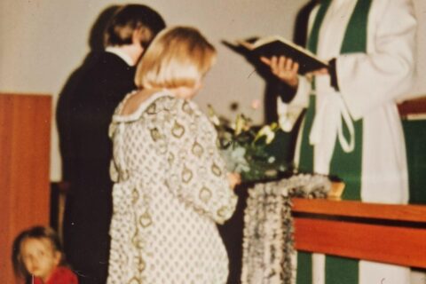 1970-luvun hääpari alttarilla papin edessä. Parin takaa kurkistaa pikkutyttä kameraan.